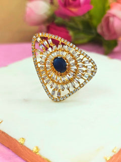 The Ferris Ring is a gold triangular adjustable ring featuring a central navy blue gemstone, surrounded by rows of sparkling American Diamonds, displayed on white marble with pink flowers in the background. Perfect American Diamond cocktail ring.