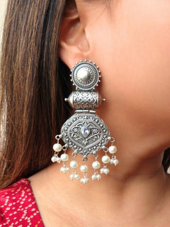 A close-up of a womans ear showcases Yamuna Earrings—ornate oxidized silver earrings with intricate designs, a central kundan stone in pear shape, and pearl dangles—ideal for weddings. She has straight brown hair and wears a red patterned top.