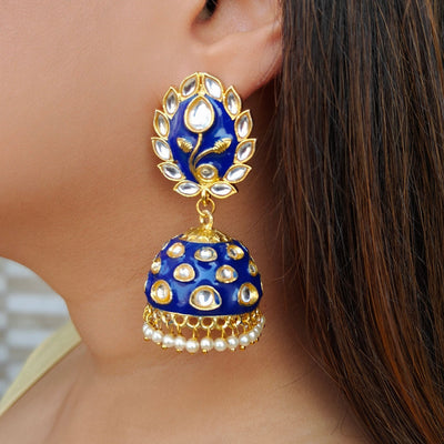 Close-up of a woman wearing a large, blue and gold traditional jhumka earring adorned with white stones and pearl beads, with a floral design on the top part.