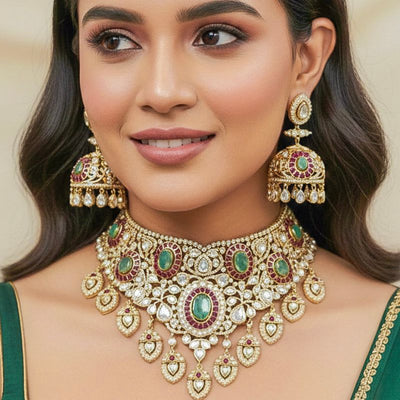 A woman wearing a green blouse, ornate gold choker necklace with green and red gemstones, matching earrings, and subtle makeup, smiling softly.