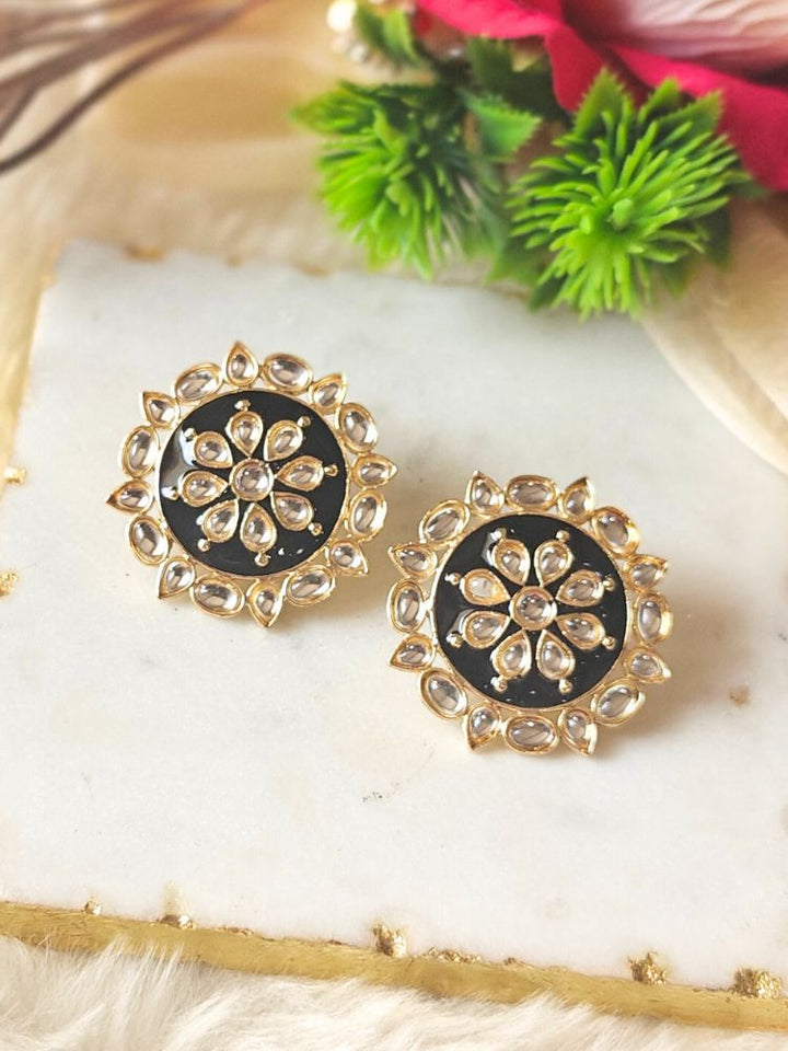The Audrey Black stud Earrings are round, gold-toned Kundan earrings, shown on a white marble slab with green leaves and red flowers in the background.