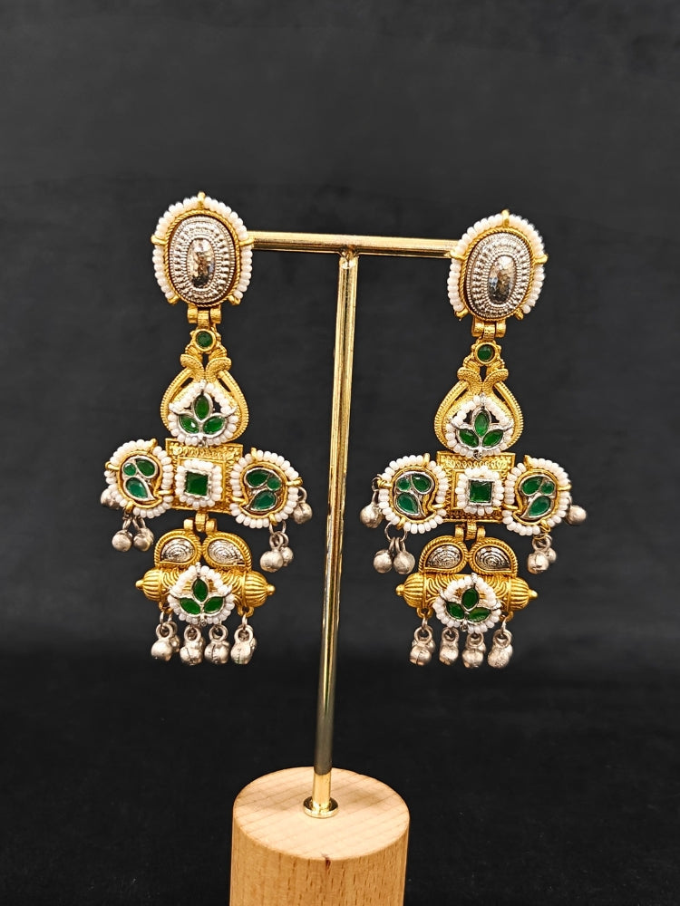 A close-up of Zaynika Green Earrings—large, ornate brass earrings with intricate detailing, white and Green stones, and hanging silver beads. The person has long dark hair and is in a dark blue patterned top.  Long Oxidized earrings, german silver earrings, navratri earrings.  Cross shaped earrings. 