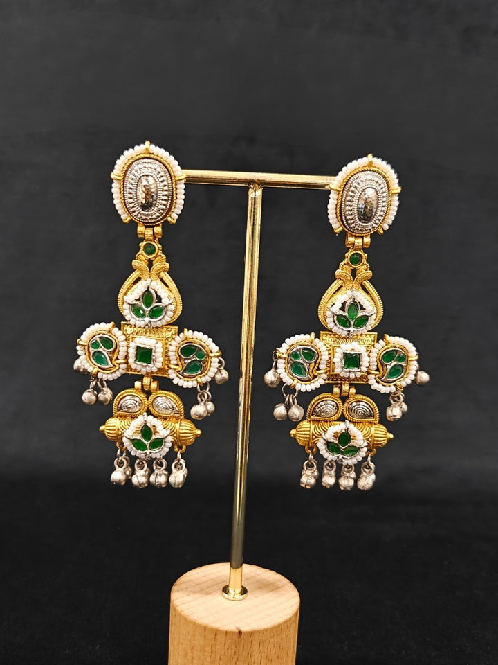 A close-up of Zaynika Green Earrings—large, ornate brass earrings with intricate detailing, white and Green stones, and hanging silver beads. The person has long dark hair and is in a dark blue patterned top.  Long Oxidized earrings, german silver earrings, navratri earrings.  Cross shaped earrings. 