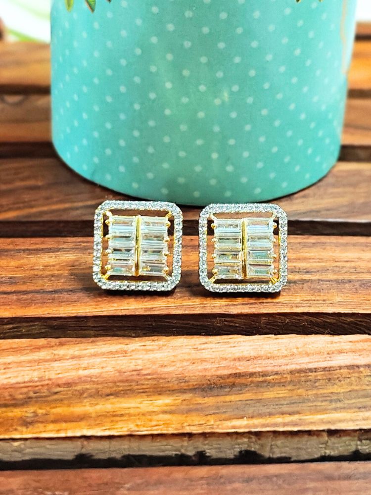 The Goldie Golden Studs are square-shaped earrings with American Diamonds in a baguette pattern, displayed on wood before a turquoise polka dot container for a chic look.