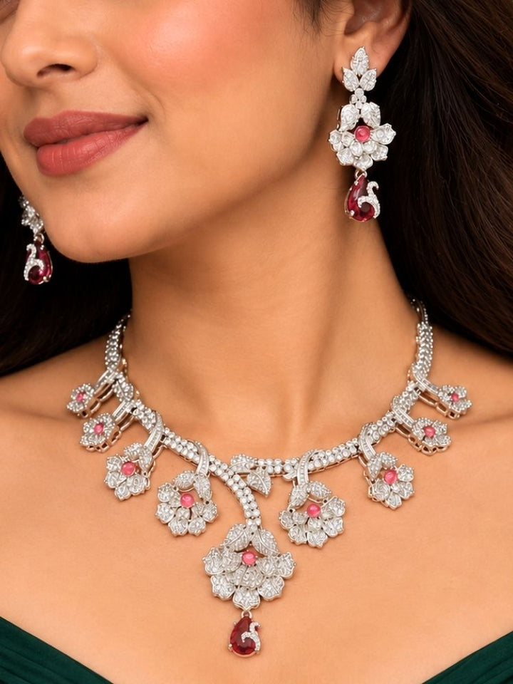 Oleander American Diamond Red Silver Tone Necklace Set – woman wearing a silver-tone American diamond floral necklace with ruby red drops and matching earrings, styled with a green outfit, artificial jewellery for cocktail parties, engagement and reception
