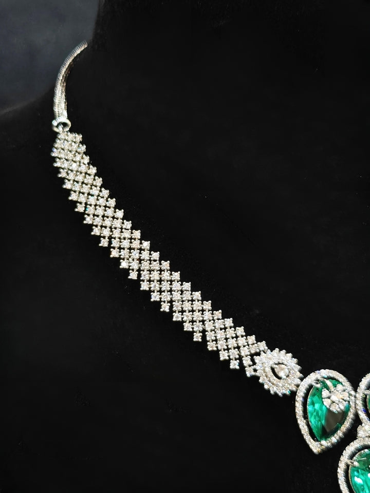 Close-up of an ornate Western (Silver Polish) Diamond Green Collar jewellery set and earrings with American Diamond and Green Colored Stones. Close-up shot of a high-quality American diamond artificial necklace worn for festive celebrations. Party wear outfit completed with a lightweight American diamond necklace.