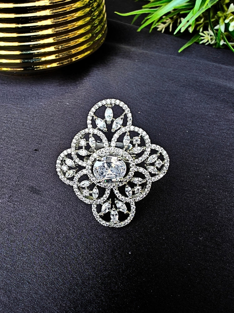 The Adeline Ring, featuring intricate symmetrical patterns and sparkling American diamonds in a silver finish, is displayed on a black surface with a gold-striped accent and green leaves in the background—perfect for wedding ceremonies.