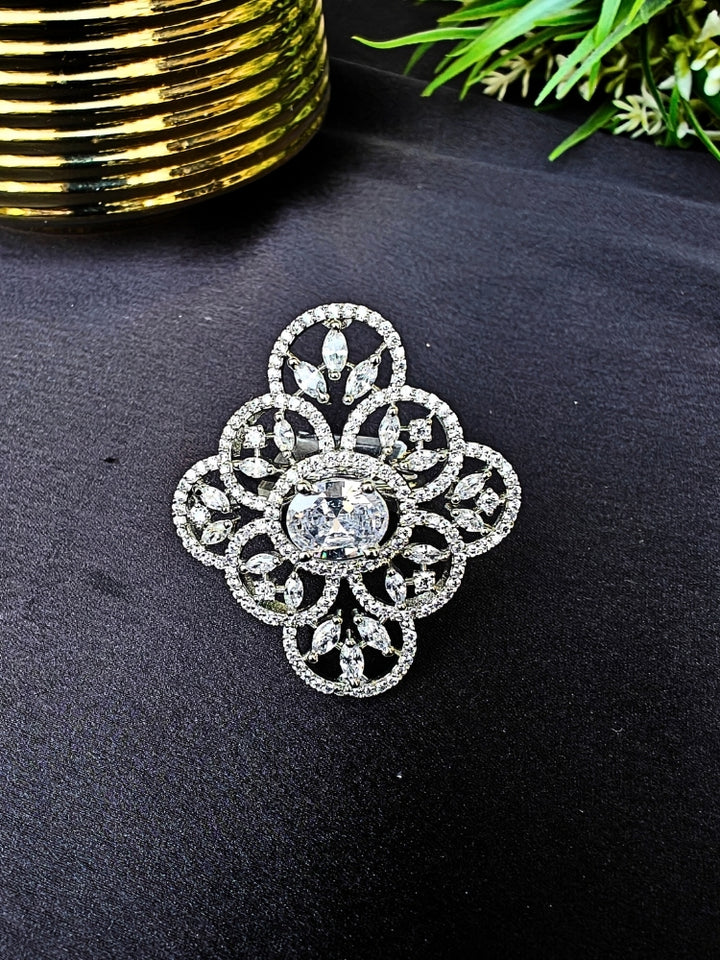 The Adeline Ring, featuring intricate symmetrical patterns and sparkling American diamonds in a silver finish, is displayed on a black surface with a gold-striped accent and green leaves in the background—perfect for wedding ceremonies.