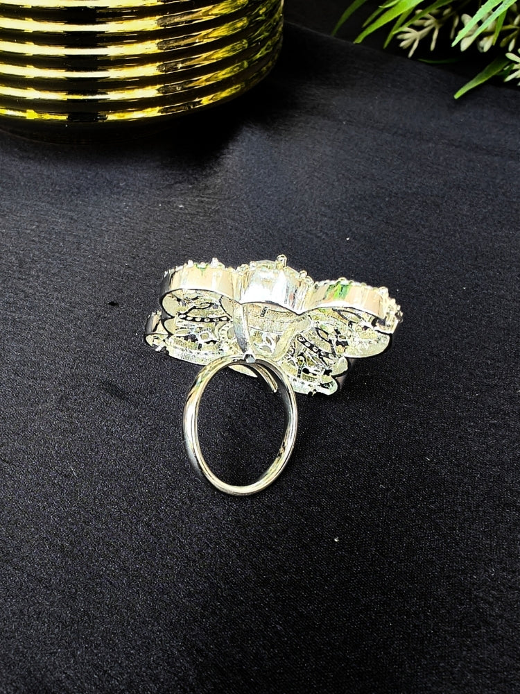 The Adeline Ring, featuring intricate symmetrical patterns and sparkling American diamonds in a silver finish, is displayed on a black surface with a gold-striped accent and green leaves in the background—perfect for wedding ceremonies.
