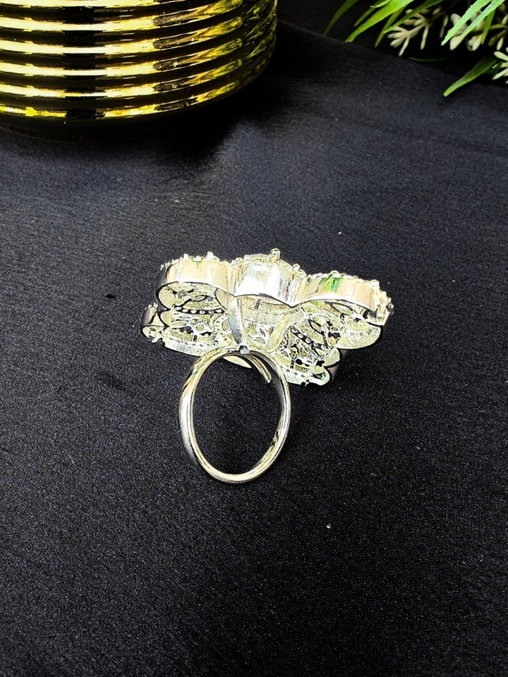 The Adeline Ring, featuring intricate symmetrical patterns and sparkling American diamonds in a silver finish, is displayed on a black surface with a gold-striped accent and green leaves in the background—perfect for wedding ceremonies.