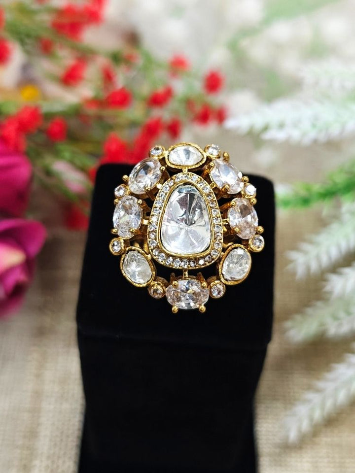 The White Adhira Ring is an ornate gold adjustable ring with a large Tyani Kundan in the middle surrounded by american diamonds, with smaller tyani kundans on the side, and oval White colored stones along with small american diamonds in the middle—displayed on black with a floral backdrop, perfect for accessorizing your wedding ceremony dress.