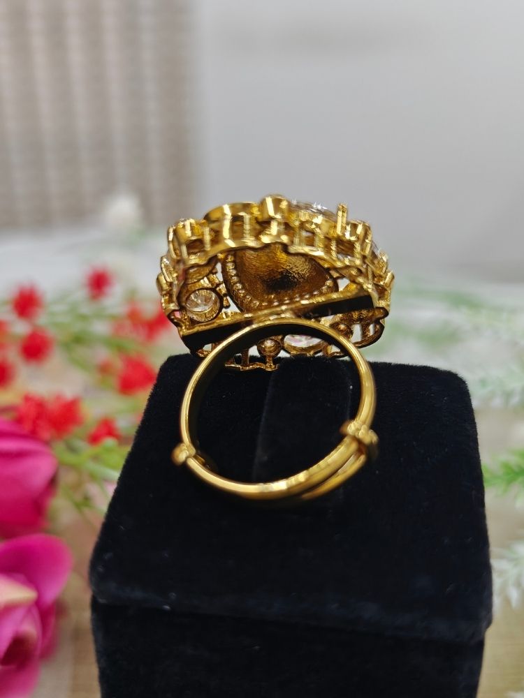 The White Adhira Ring is an ornate gold adjustable ring with a large Tyani Kundan in the middle surrounded by american diamonds, with smaller tyani kundans on the side, and oval White colored stones along with small american diamonds in the middle—displayed on black with a floral backdrop, perfect for accessorizing your wedding ceremony dress.