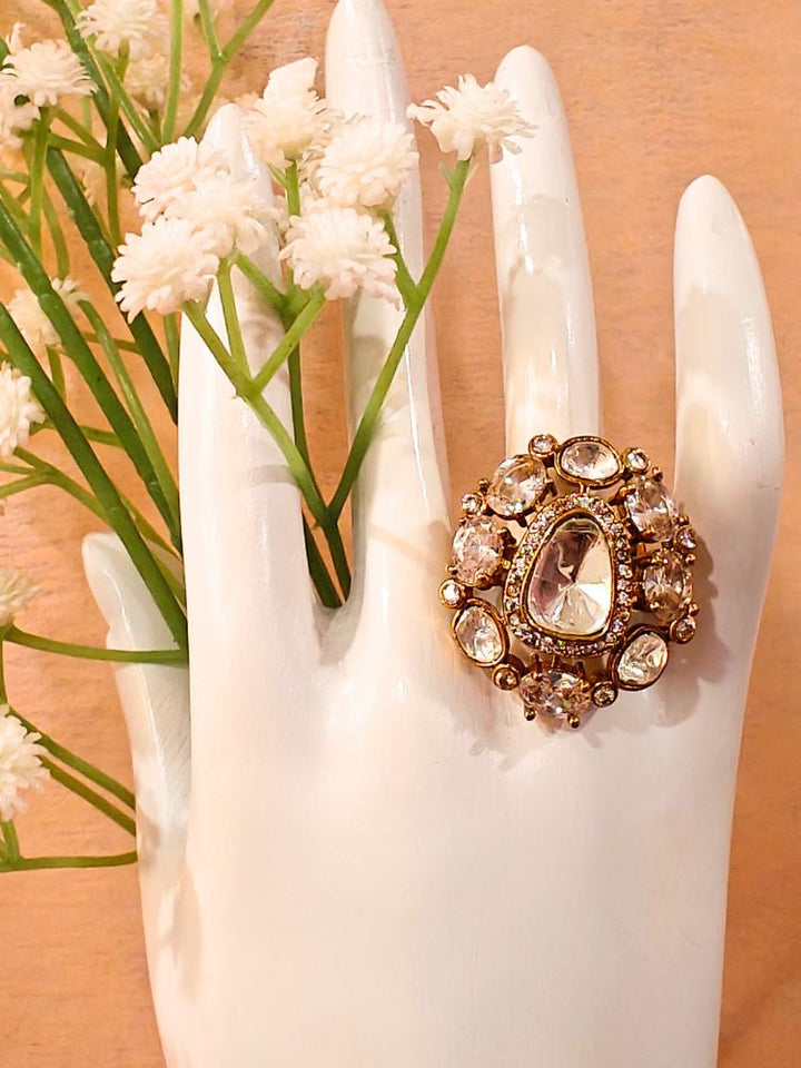 The White Adhira Ring is an ornate gold adjustable ring with a large Tyani Kundan in the middle surrounded by american diamonds, with smaller tyani kundans on the side, and oval White colored stones along with small american diamonds in the middle—displayed on White with White Flowers, perfect for accessorizing your wedding ceremony dress.