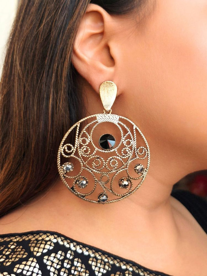 A close-up of a woman with long brown hair in a netted-patterned top, wearing Aditi Earrings—large, round alloy filigree earrings with intricate swirls, embedded black crystal stones, and a central black gem.