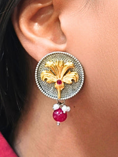 A close-up of a womans ear wearing Advika Pink Earrings: large round brass earrings with gold floral design, silver textured background, center red gemstone, and dangling red and white beads.