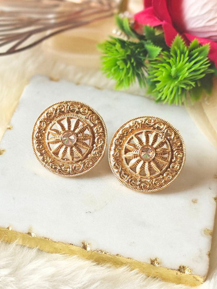 The Akhila Earrings are round gold stud earrings featuring a wheel shape design, with a central gemstone and Reverse American Diamond accents, displayed on a marble slab with artificial green and red flowers in the background.