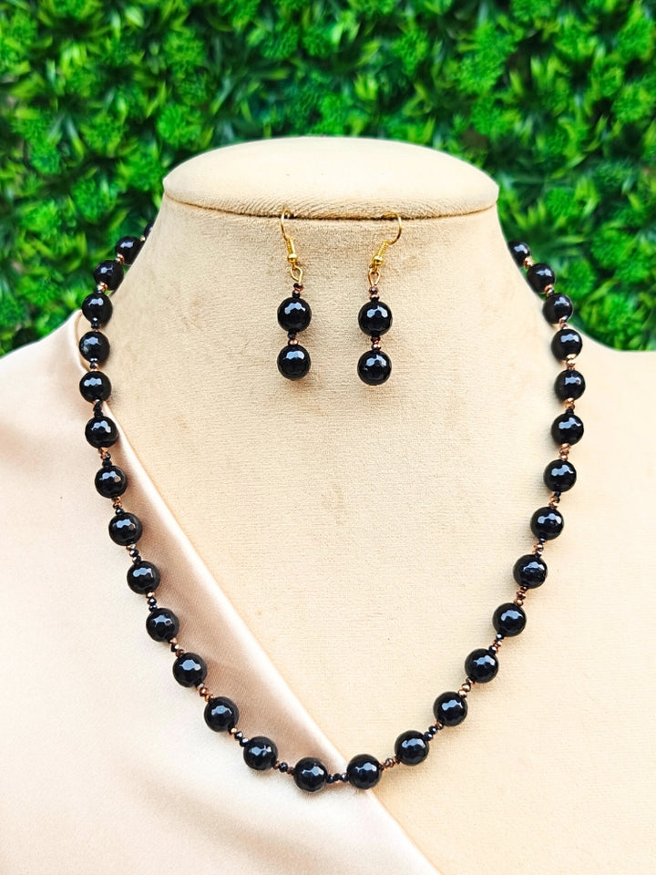 The Alberta Necklace Set features a black beaded necklace and matching earrings with gold hooks, showcased on a beige mannequin against a vibrant green leafy background.