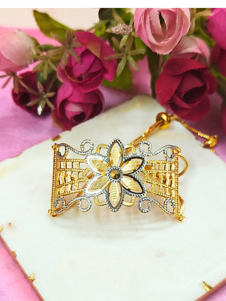 The Alisha Gold Bracelet, featuring adjustable fit and intricate filigree with geometric patterns, is displayed on a white marble slab amid pink and red artificial flowers against a pink background—ideal for weddings.
