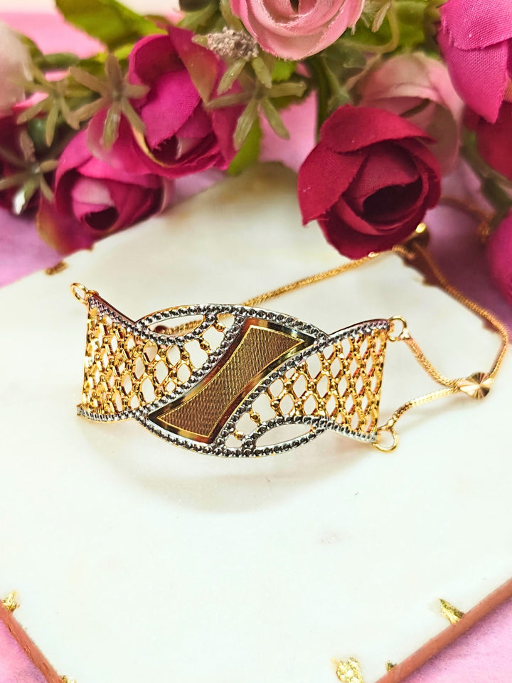 The Alisha Gold Bracelet, featuring adjustable fit and intricate filigree with geometric patterns, is displayed on a white marble slab amid pink and red artificial flowers against a pink background—ideal for weddings.