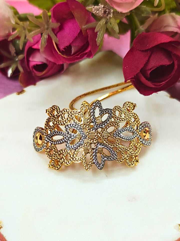 The Alisha Gold Bracelet, featuring adjustable fit and intricate filigree with geometric patterns, is displayed on a white marble slab amid pink and red artificial flowers against a pink background—ideal for weddings.