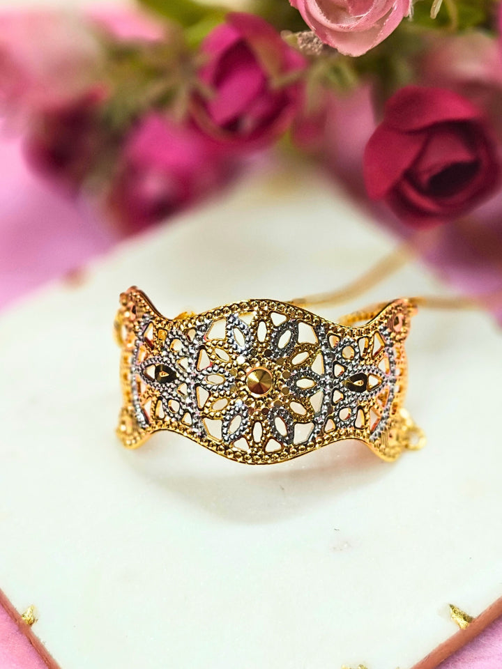 The Alisha Gold Bracelet, featuring adjustable fit and intricate filigree with geometric patterns, is displayed on a white marble slab amid pink and red artificial flowers against a pink background—ideal for weddings.