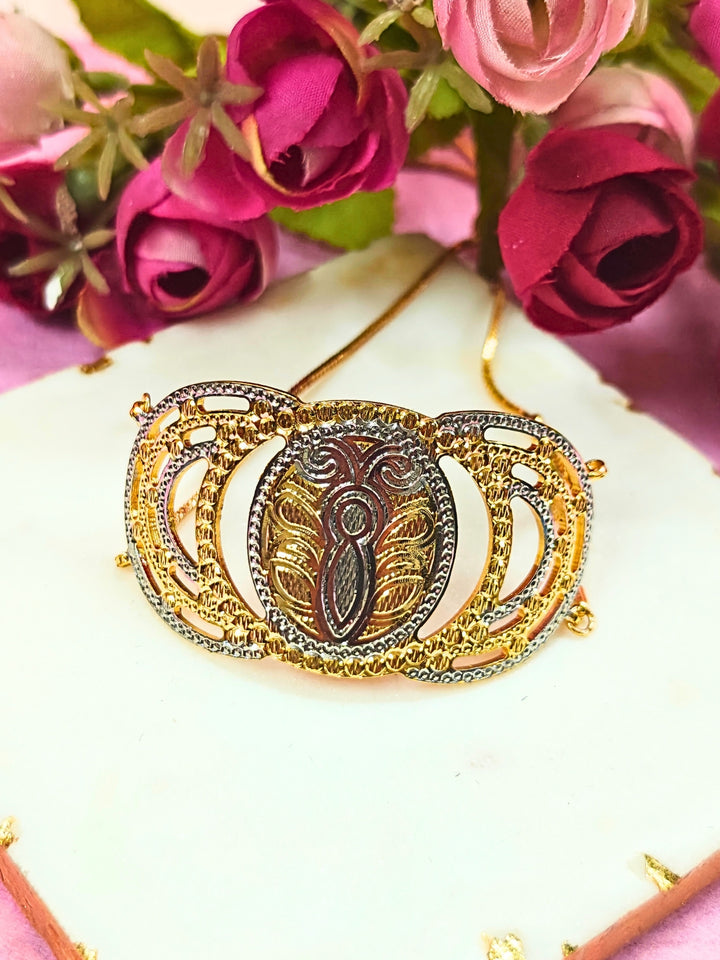 The Alisha Gold Bracelet, featuring adjustable fit and intricate filigree with geometric patterns, is displayed on a white marble slab amid pink and red artificial flowers against a pink background—ideal for weddings.
