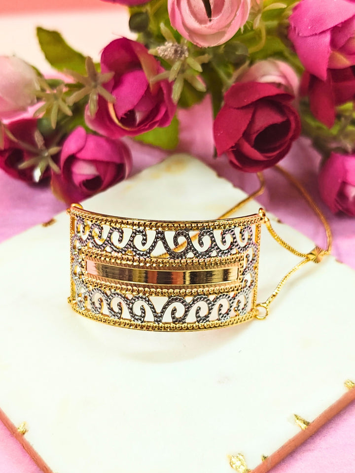 The Alisha Gold Bracelet, featuring adjustable fit and intricate filigree with geometric patterns, is displayed on a white marble slab amid pink and red artificial flowers against a pink background—ideal for weddings.