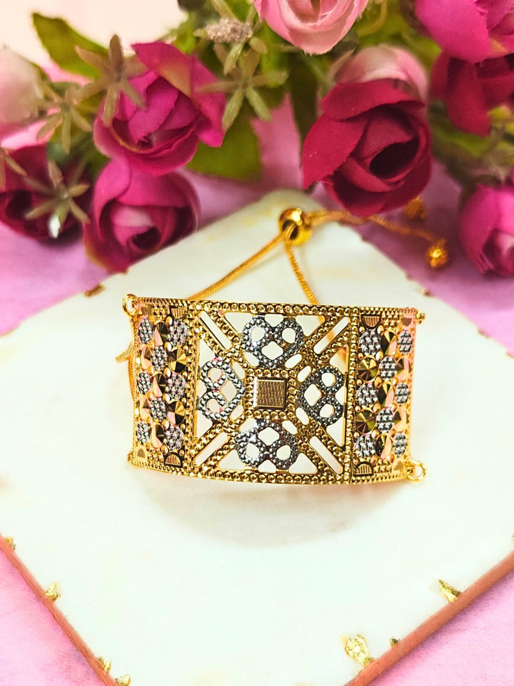 The Alisha Gold Bracelet, featuring adjustable fit and intricate filigree with geometric patterns, is displayed on a white marble slab amid pink and red artificial flowers against a pink background—ideal for weddings.