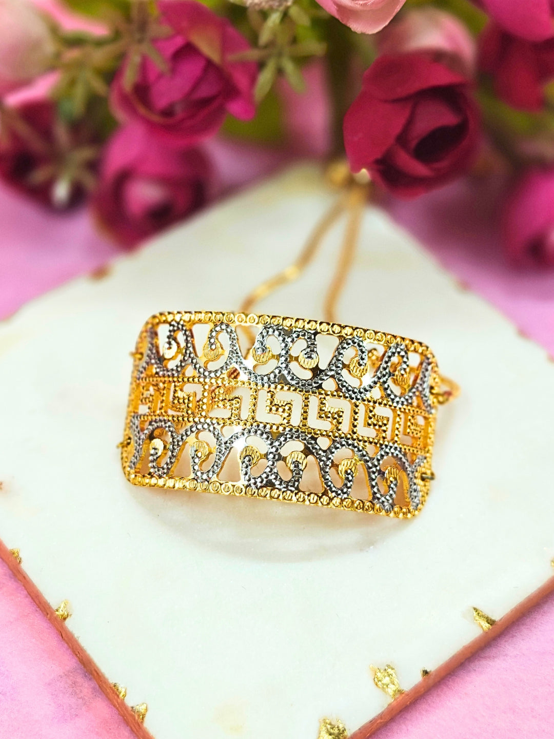 The Alisha Gold Bracelet, featuring adjustable fit and intricate filigree with geometric patterns, is displayed on a white marble slab amid pink and red artificial flowers against a pink background—ideal for weddings.