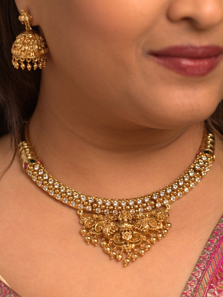 A woman wearing the Ambhira Laxmi Hasli Necklace Set featuring a traditional gold Collar Hasli necklace and matching earrings with Goddess Laxmi in the middle, along with American Diamonds and Colored Stones. Wedding guest look completed with a Hasli temple necklace set.