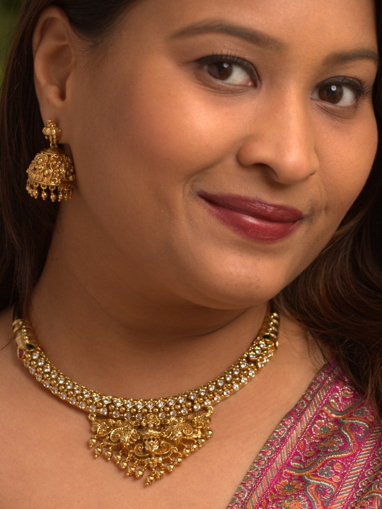 A woman wearing the Ambhira Laxmi Hasli Necklace Set featuring a traditional gold Collar Hasli necklace and matching earrings with Goddess Laxmi in the middle, along with American Diamonds and Colored Stones. Wedding guest look completed with a Hasli temple necklace set.