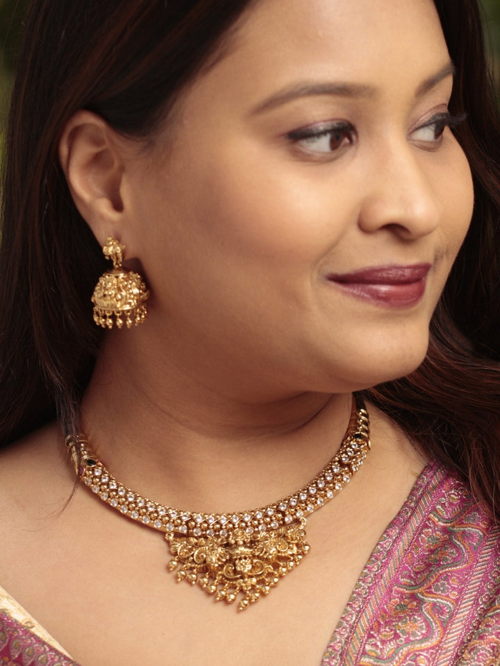 A woman wearing the Ambhira Laxmi Hasli Necklace Set featuring a traditional gold Collar Hasli necklace and matching earrings with Goddess Laxmi in the middle, along with American Diamonds and Colored Stones. Wedding guest look completed with a Hasli temple necklace set.