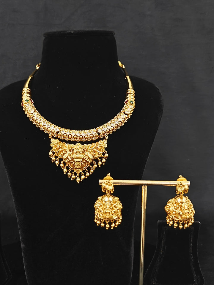 The Ambhira Laxmi Hasli Necklace Set featuring a traditional gold Collar Hasli necklace and matching earrings with Goddess Laxmi in the middle, along with American Diamonds and Colored Stones. Wedding guest look completed with a Hasli temple necklace set.