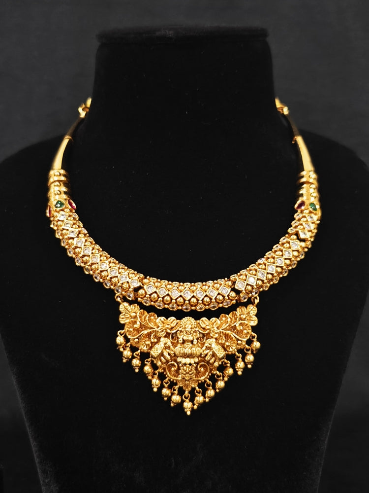 The Ambhira Laxmi Hasli Necklace Set featuring a traditional gold Collar Hasli necklace and matching earrings with Goddess Laxmi in the middle, along with American Diamonds and Colored Stones. Wedding guest look completed with a Hasli temple necklace set.