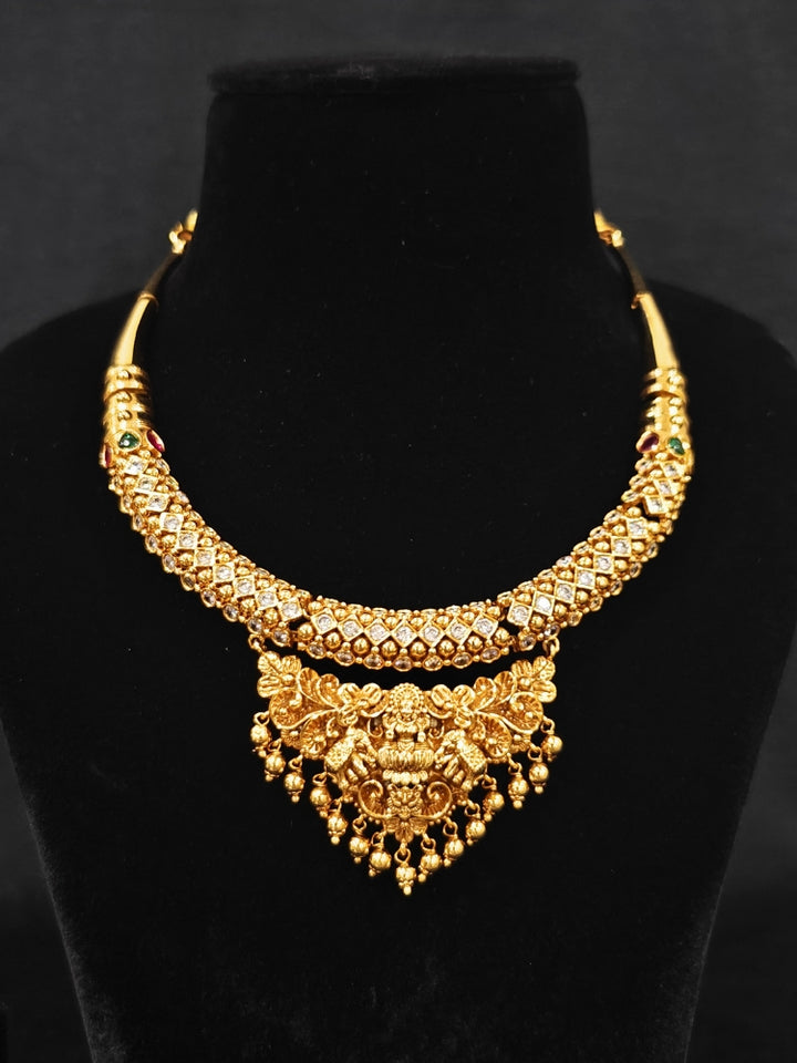 The Ambhira Laxmi Hasli Necklace Set featuring a traditional gold Collar Hasli necklace and matching earrings with Goddess Laxmi in the middle, along with American Diamonds and Colored Stones. Wedding guest look completed with a Hasli temple necklace set.