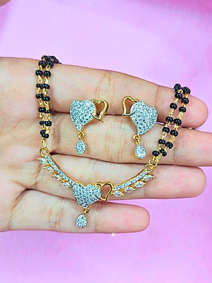 A hand holds the Ambi Mangalsutra—a gold necklace with black mangalsutra beads and matching earrings, pendant featuring sparkling American Diamonds—set against a soft pink background, perfect for your wedding ceremony dress. Diamond mangalsutra. Modern mangalsutra. 