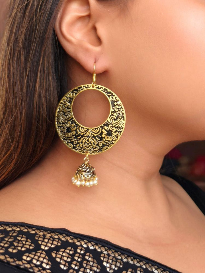 A close-up of a womans ear adorned with Amyra Earrings—large, round, gold-toned Meenakari earrings featuring intricate floral designs, along with a small jhumki at the bottom with hanging pearls. Her long brown hair and black outfit make them perfect for a wedding ceremony.