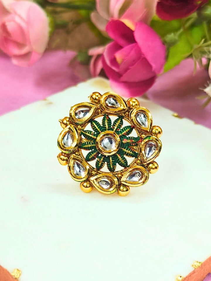 The Anamika Dark Green Kundan Ring is a round, ornate gold adjustable ring with teardrop-shaped white Kundan stones and Dark Green enamel, shown upright on a white marble slab with blurred pink and red flowers in the background.