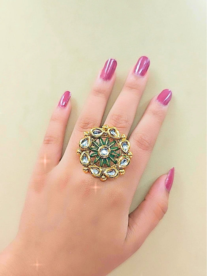 The Anamika Dark Green Kundan Ring is a round, ornate gold adjustable ring with teardrop-shaped white Kundan stones and dark green enamel, shown on a girls hand with pink nail paint. 