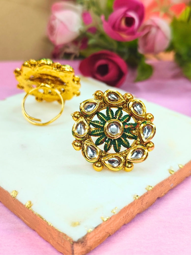 The Anamika Dark Green Kundan Ring is a round, ornate gold adjustable ring with teardrop-shaped white Kundan stones and Dark Green enamel, shown upright on a white marble slab with blurred pink and red flowers in the background.