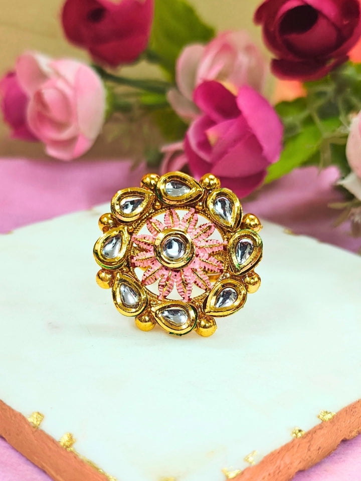 The Anamika Mint Pink Kundan Ring is a round, ornate gold adjustable ring with teardrop-shaped white Kundan stones and mint Pink enamel, shown upright on a white marble slab with blurred pink and red flowers in the background.