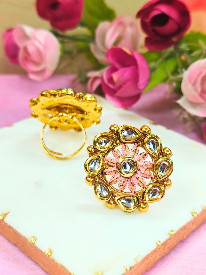 The Anamika Mint Pink Kundan Ring is a round, ornate gold adjustable ring with teardrop-shaped white Kundan stones and mint Pink enamel, shown upright on a white marble slab with blurred pink and red flowers in the background.