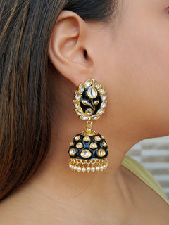A close-up of a womans ear wearing Ananya Black Kundan Jhumkas, featuring Black and gold enamel, teardrop-shaped white stones, and pearl details, set against her straight brown hair.