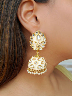 A close-up of a womans ear wearing Ananya Ivory Kundan Jhumkas, featuring Ivory and gold enamel, teardrop-shaped white stones, and pearl details, set against her straight brown hair.