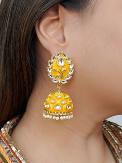 A close-up of a womans ear wearing Ananya Mustard Yellow Kundan Jhumkas, featuring Mustard Yellow and gold enamel, teardrop-shaped white stones, and pearl details, set against her straight brown hair.