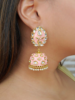 A close-up of a womans ear wearing Ananya Peach Kundan Jhumkas, featuring Peach and gold enamel, teardrop-shaped white stones, and pearl details, set against her straight brown hair.