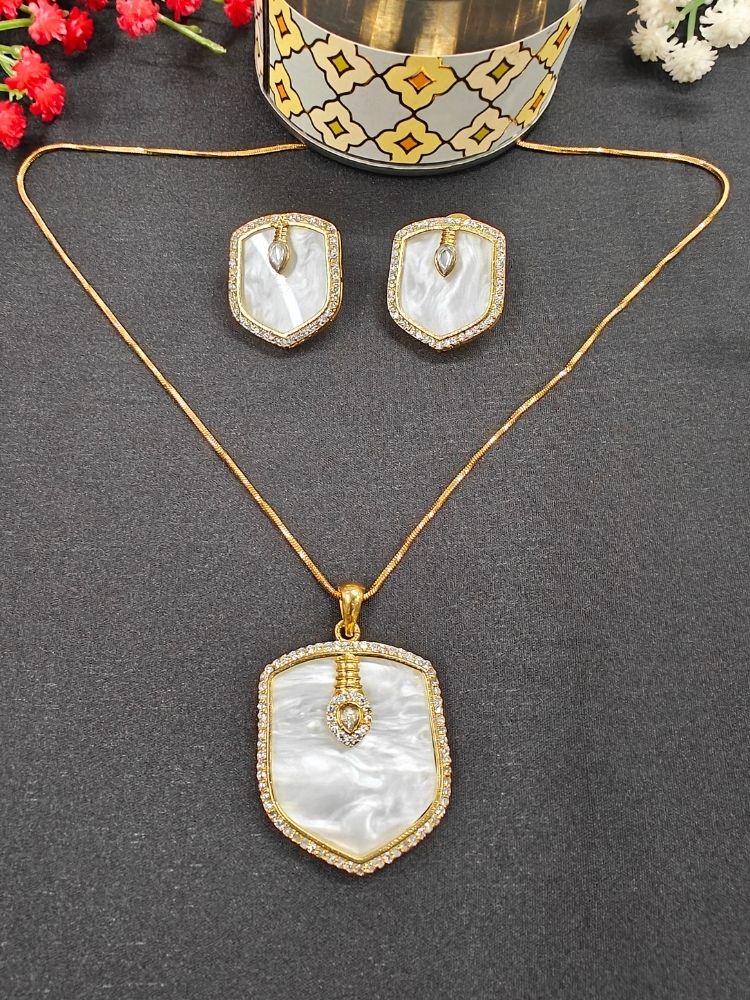 The Anchor Pendant Set is a gold necklace and earring set with shield-shaped or anchor shaped white pendants, mother-of-pearl centers, and sparkling American Diamonds, shown on a black surface with a decorative container and red flowers.