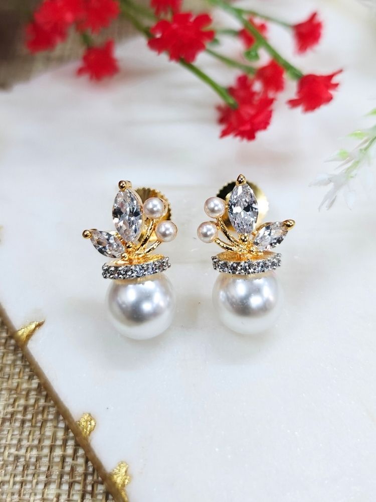 The Andie stud Earrings feature elegant gold-plated brass earrings, a large round pearl at the bottom, marquise-cut American Diamonds, crystal accents, and tiny pearls, beautifully displayed on a white surface with red flower buds in the background. 
