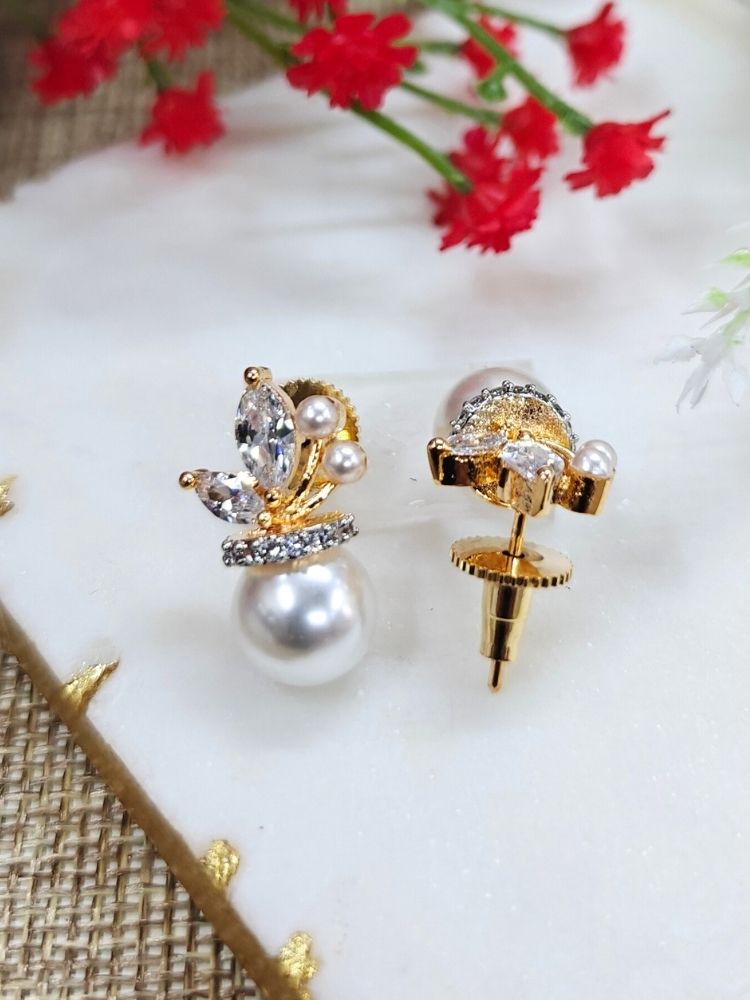 The Andie stud Earrings feature elegant gold-plated brass earrings, a large round pearl at the bottom, marquise-cut American Diamonds, crystal accents, and tiny pearls, beautifully displayed on a white surface with red flower buds in the background. 