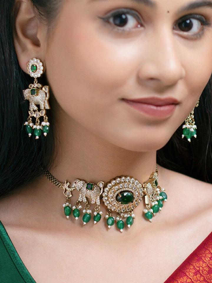 A woman wearing the Aneya Dark Green Choker Necklace Set, featuring ornate gold-toned jewelry with Dark Green and white colored stones and elephant-shaped motifs in the necklace and earrings, poses in traditional attire, her long dark hair partially visible.